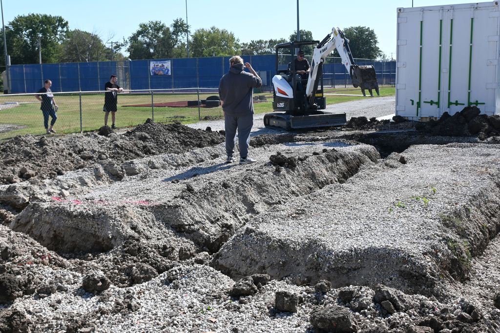 As students look on, Mr. Gilpin operates a mini excavator to dig out the footings for the greenhouse.