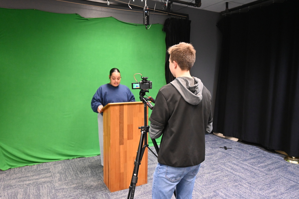 One student stands behing the podium to help  another adjust camera angles.