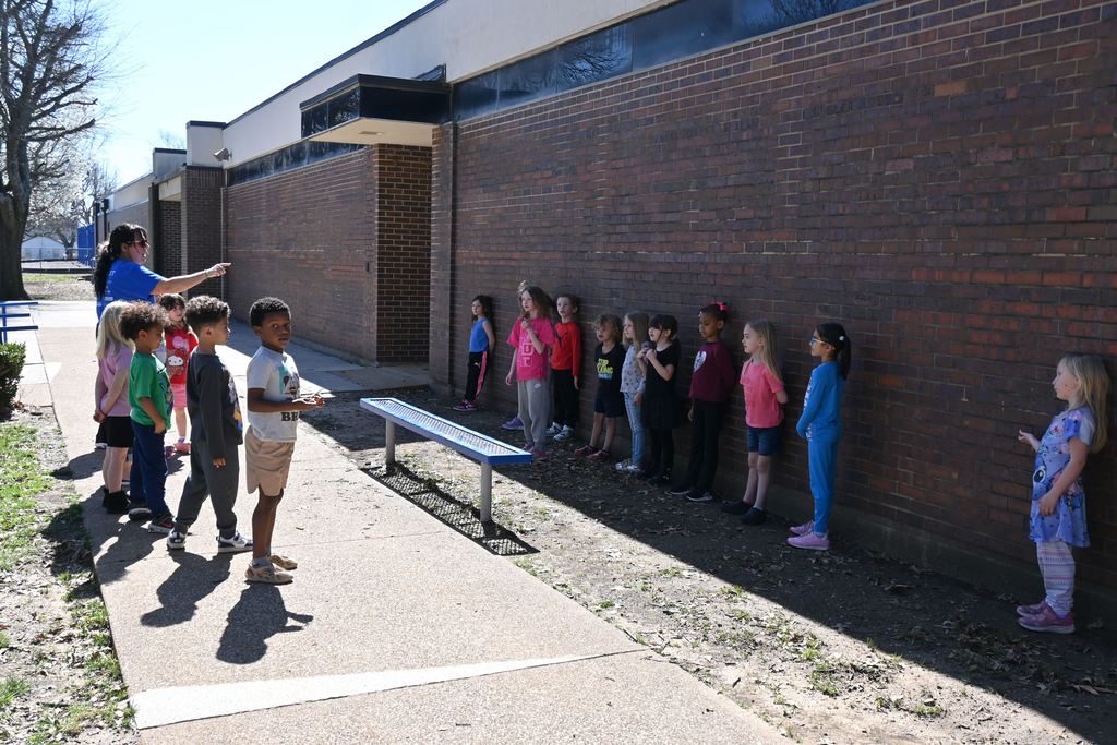 Some students stand in the shade of the building and others in the sun.