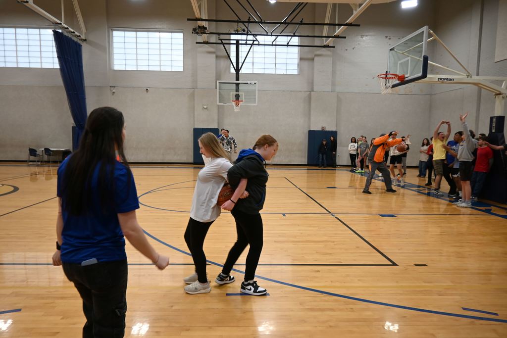 Two girls stand back to back with a ball between them, and race for the finish line.
