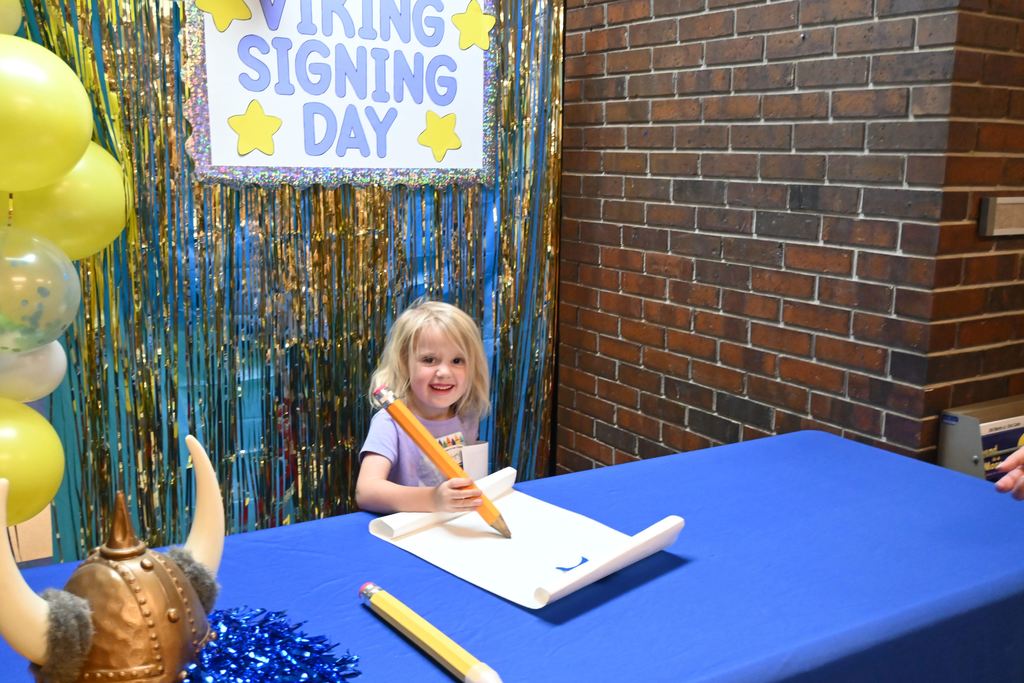 Children got their picture taken with a giant pencil signing a paper with a Viking Signing Day poster behind them