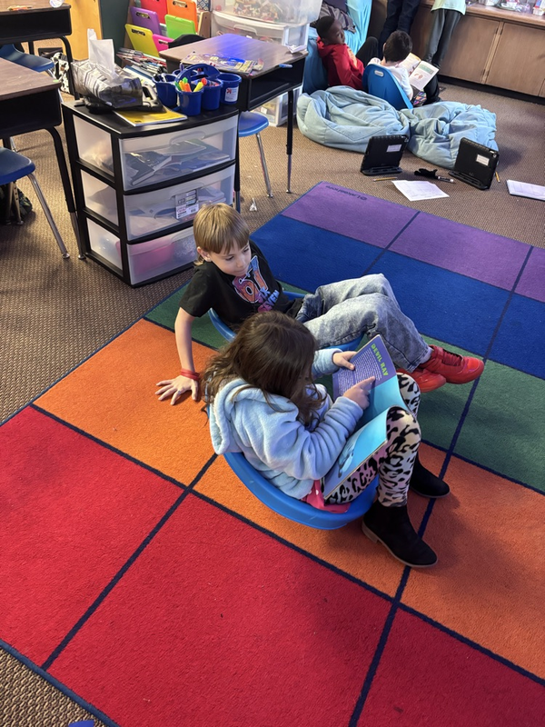 Students sitting around the room on the floor or on low slung chairs reading in pairs.