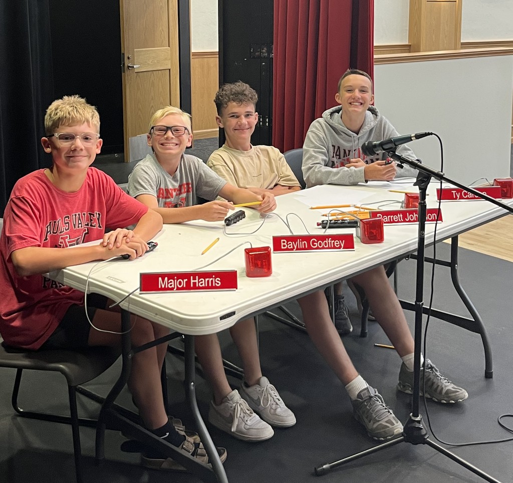 Four junior high students sit at a table with nameplates and buzzers during an academic team event.