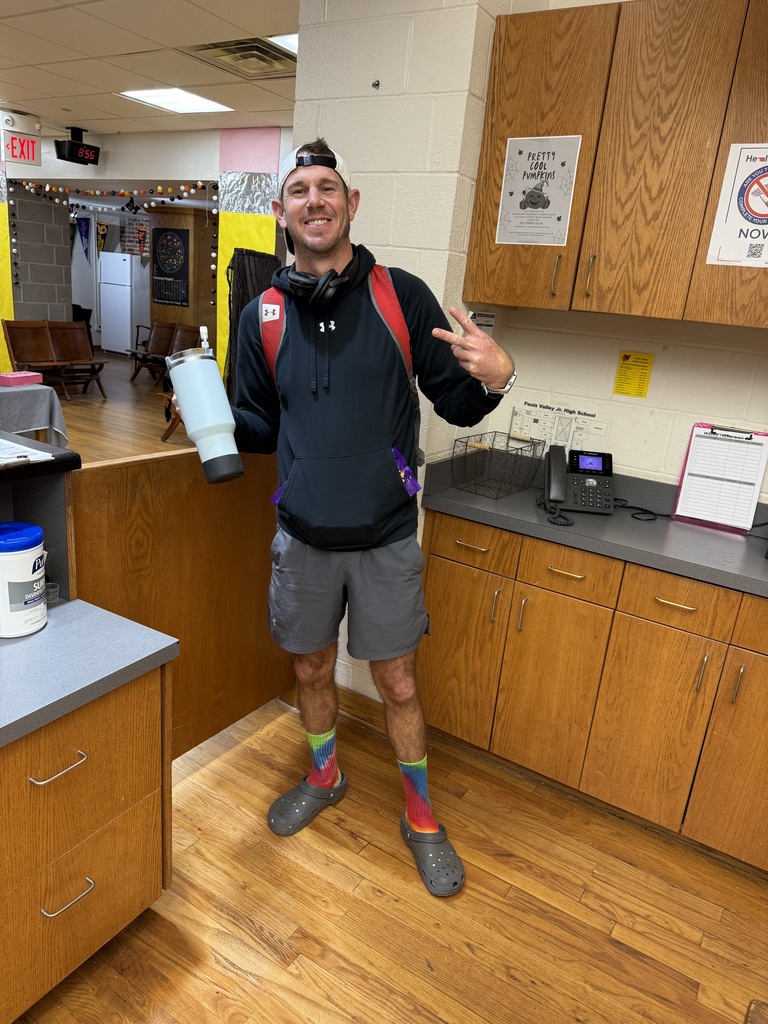 A man stands in a school office area smiling and making a peace sign with one hand while holding a large light blue tumbler in the other. He’s wearing a black hoodie, gray shorts, tie-dye socks, and gray Crocs, with a red backpack, white cap worn backward, and headphones around his neck. The background shows wooden cabinets, a countertop with cleaning supplies, and posters on the wall, with a lounge area visible through an open doorway.