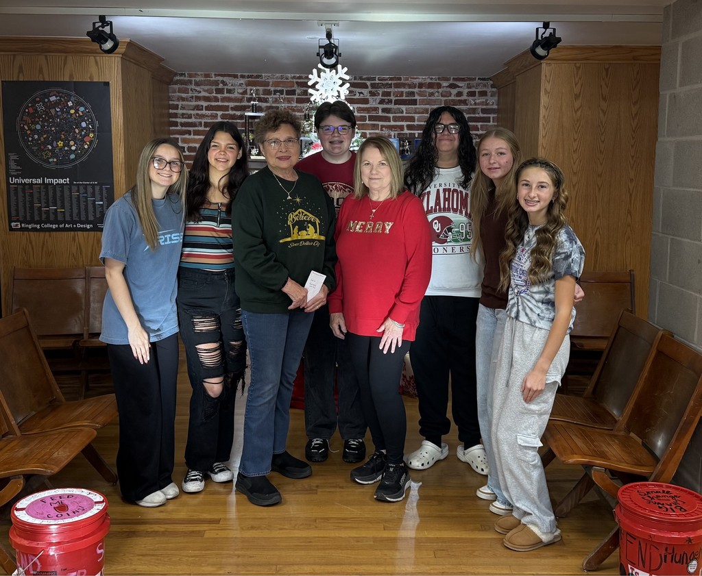 A group of nine people stand together in a cozy indoor space with wooden benches, brick walls, and holiday decorations, including a white snowflake hanging above them. They are smiling and standing close in a casual, friendly pose. The group includes teens and two adults, all dressed in casual or festive clothing—some wearing Christmas-themed shirts. Red donation buckets sit on the floor in front and to the sides.