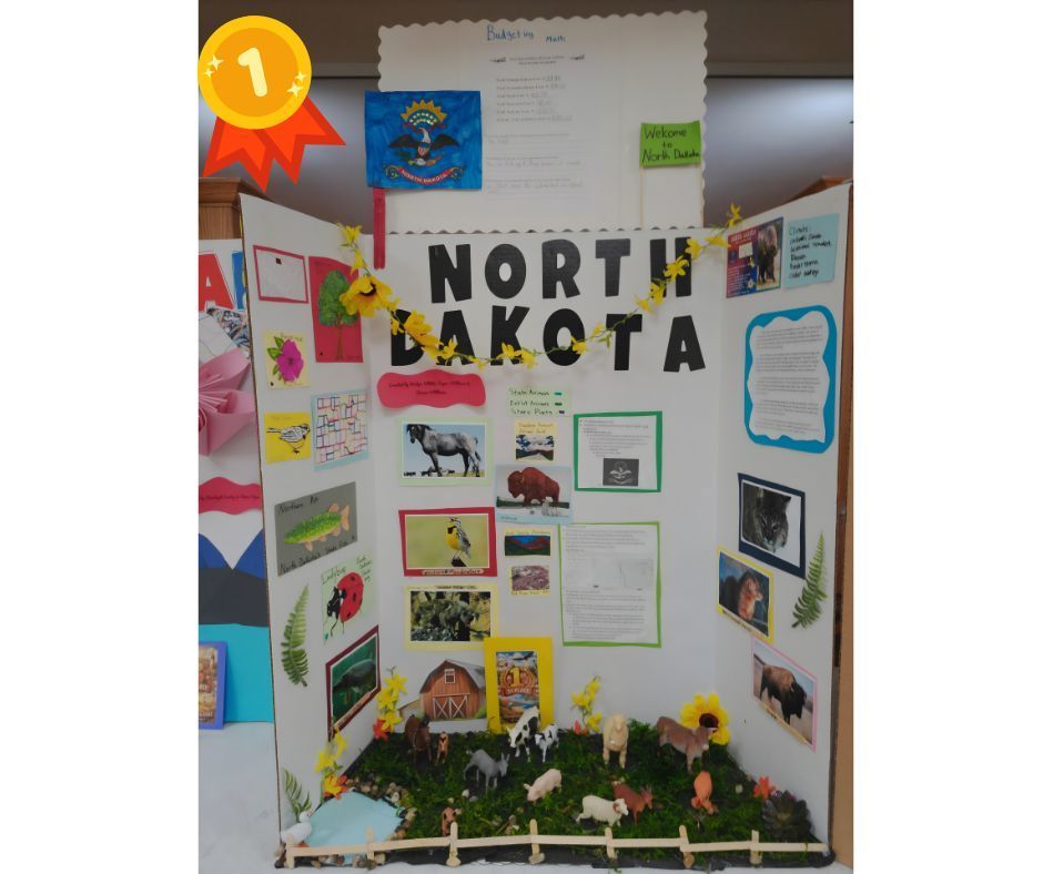 Tri-fold presentation board titled “North Dakota,” marked with a first-place ribbon icon in the top left corner. The display includes colorful text blocks, photos, and drawings about North Dakota, such as state facts, wildlife, landscapes, and attractions. A small North Dakota state flag is displayed near the top. The bottom of the board features a 3D diorama with grass, a small fence, a barn, and miniature farm animals including cows, horses, pigs, and sheep. The project is neatly arranged and visually detailed, showcasing research and creativity.