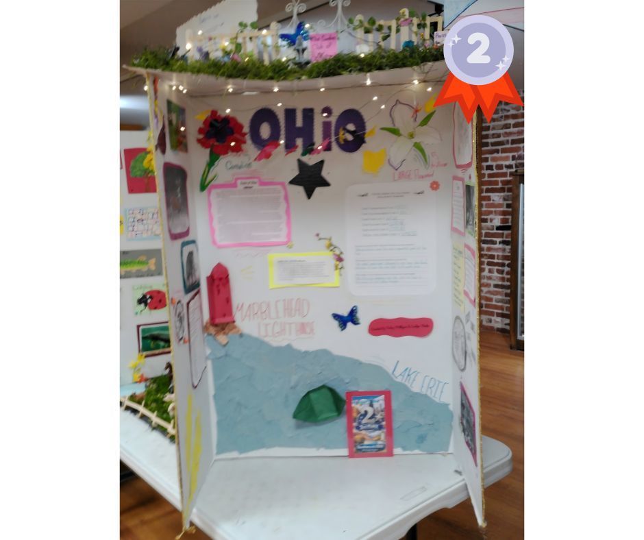 Tri-fold presentation board titled “Ohio,” marked with a second-place ribbon icon in the top right corner. The display features colorful decorations, including flowers, butterflies, stars, and drawings of the state outline. Typed and handwritten fact sheets highlight Ohio topics such as culture, landmarks, and Lake Erie. The bottom portion includes a textured blue paper design representing water and shoreline. Decorative greenery and string lights trim the top of the board, creating a bright, creative project display.