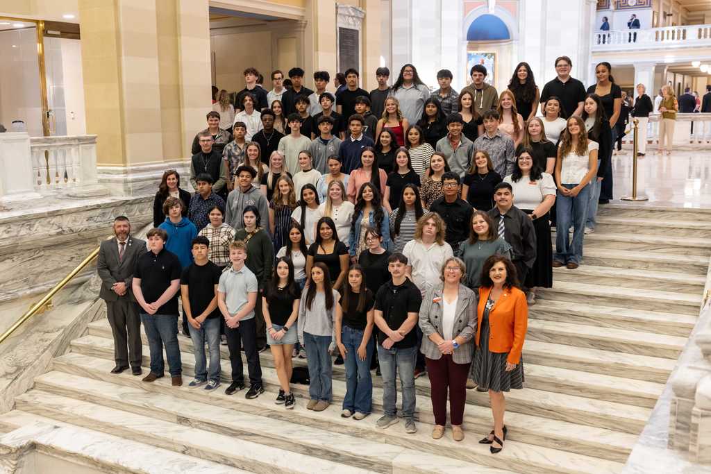 A large group of students and a few adults pose for a formal group photo on wide marble stairs inside a grand, historic building. The space features tall columns, arched doorways, and polished stone floors. The students are arranged in several rows, dressed in a mix of casual and semi-formal clothing, while a few staff members stand in front. The setting and composition suggest a school trip or special academic event, possibly at a state capitol or government building.