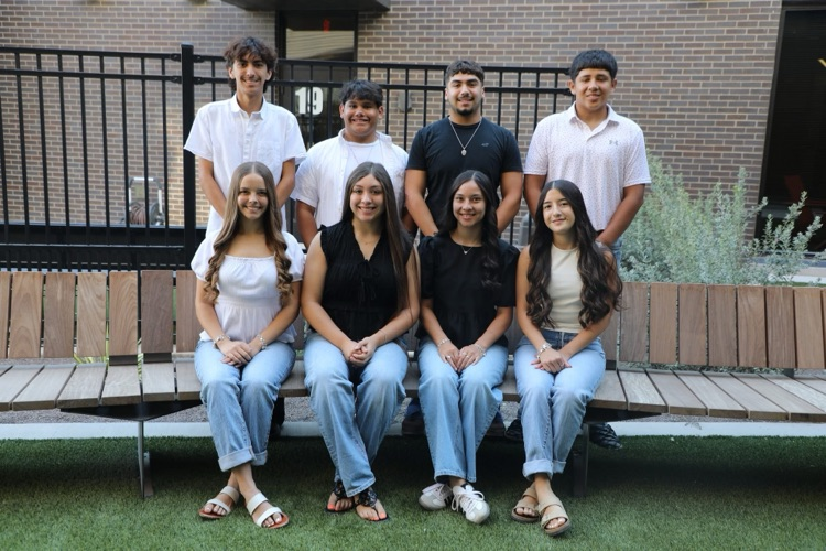 2025 Pecos Eagle Homecoming Court posing together outdoors in front of brick wall and a black metal fence. Front four girls are sitting side by side wearing light blue jeans with dressy tops. Back row boys are standing behind them, dressed causally. 