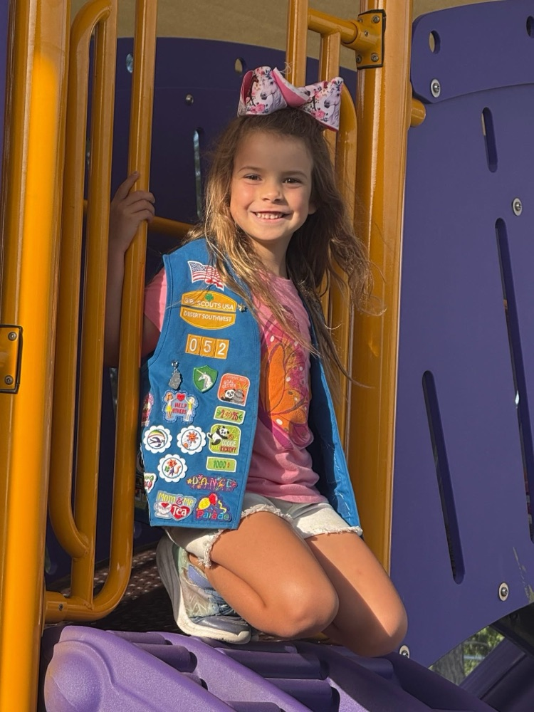 A smiling student with a blue vest with badges. She is on the playground with a pink patterned bow.