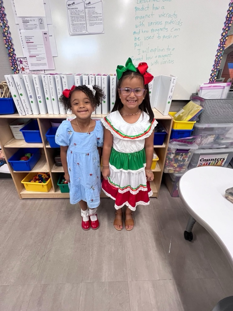 Two girl students lined up side by side in a classroom, wearing dresses that consists of blue, green, white, and red.