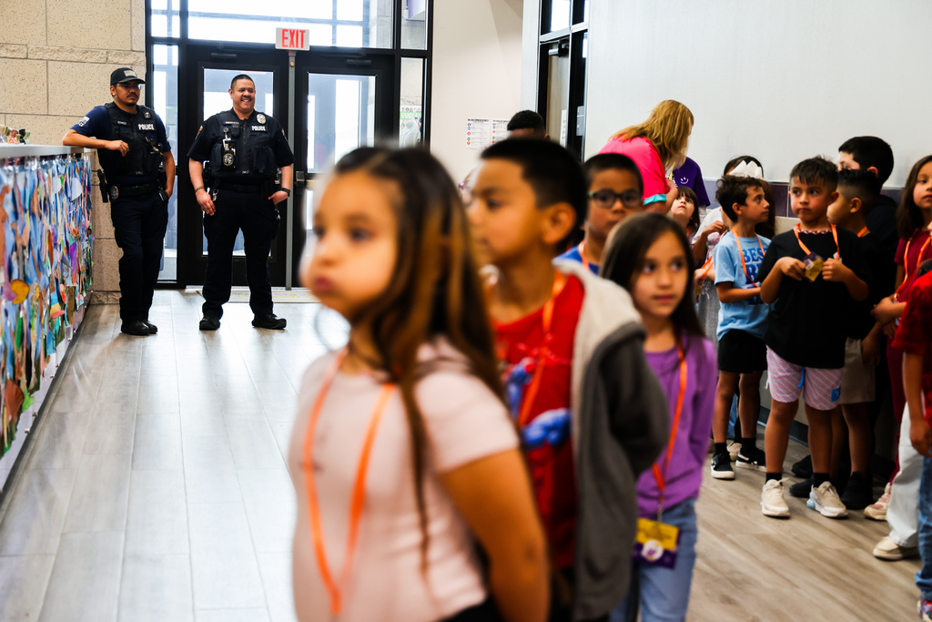 A group of young students standing in a school hallway with artwork displayed on the walls and two law enforcement officers standing nearby.