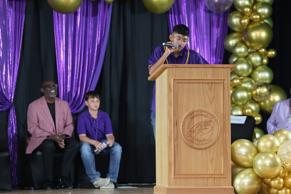 A person standing at a podium on a stage, with two other people sitting in the background. The stage is decorated with purple curtains and golden balloons.
