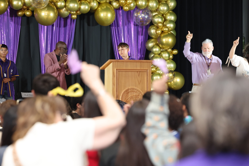 Large crowd attending an event or celebration, with balloons and decorations in gold and purple colors, and people raising their hands.