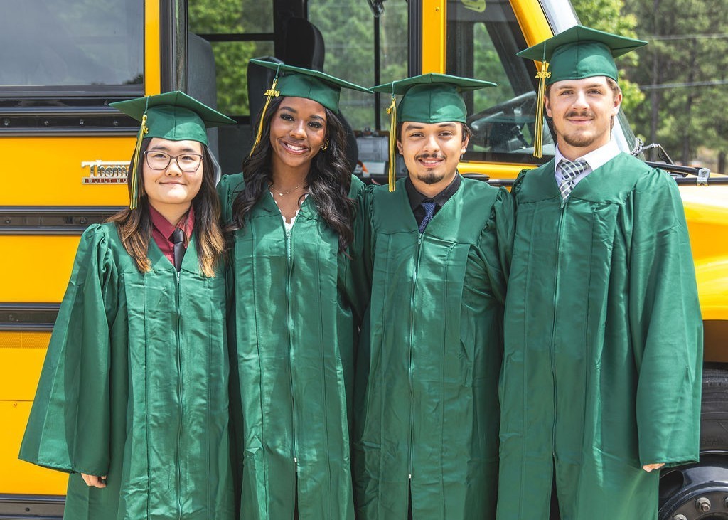 Group of senior students in green cap and gowns in front of school bus