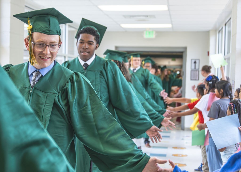 Seniors in green cap and gowns holding out hands to young students while walking in hallway