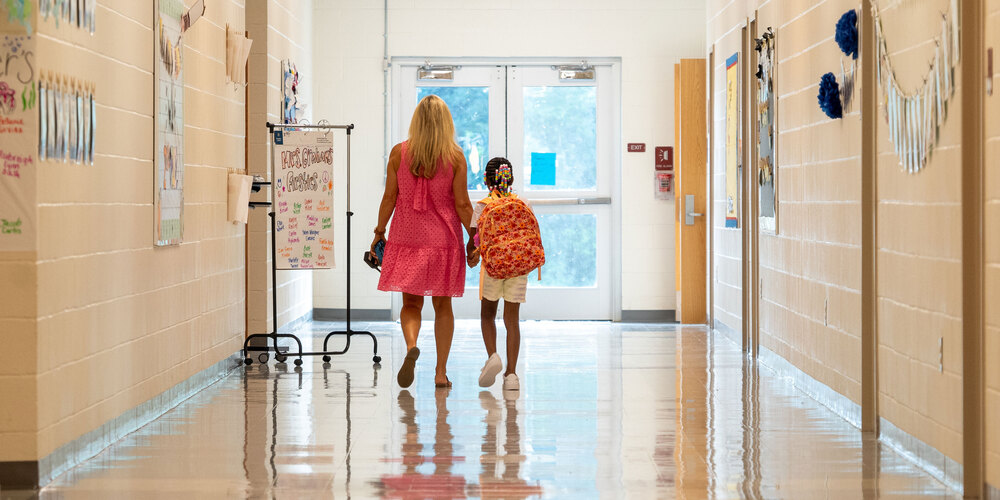 TEACHER HOLDING HANDS WITH A STUDENT WHILE WALKING DOWN A HALLWAY
