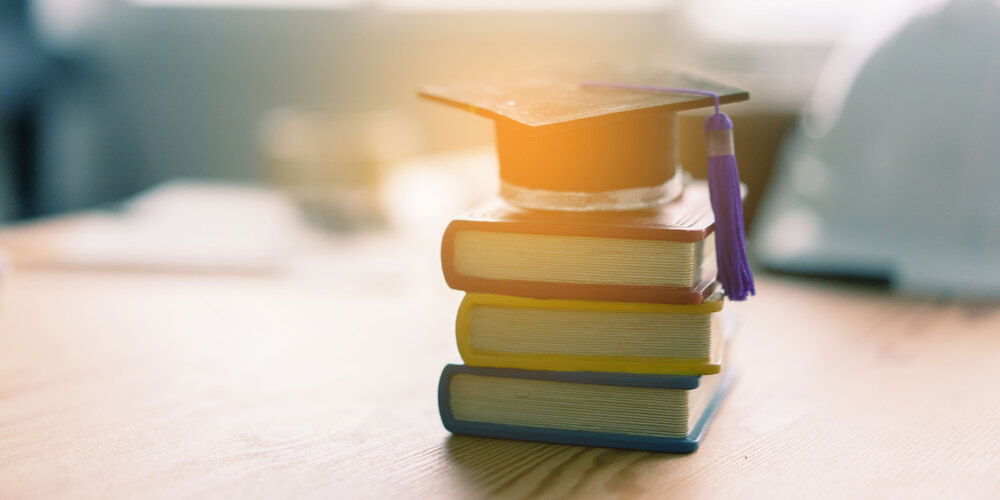 BOOKS ON A DESK WITH A GRADUATION CAP ON TOP OF THE BOOK STACK