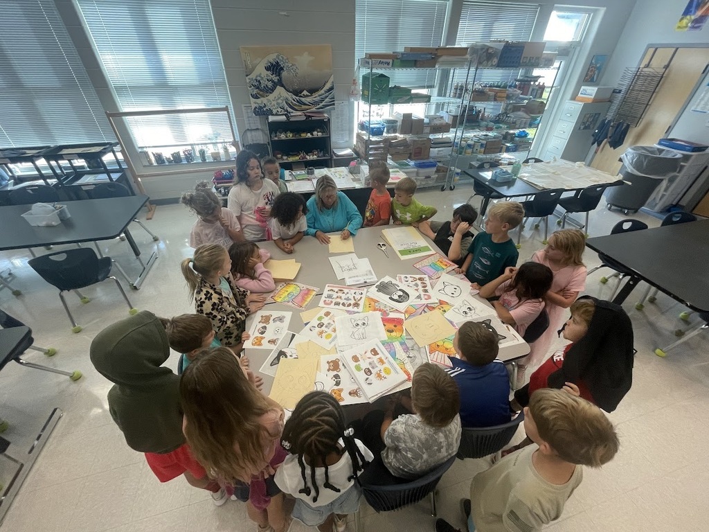 2nd grade students standing around one table watching art teacher demonstrate the lesson