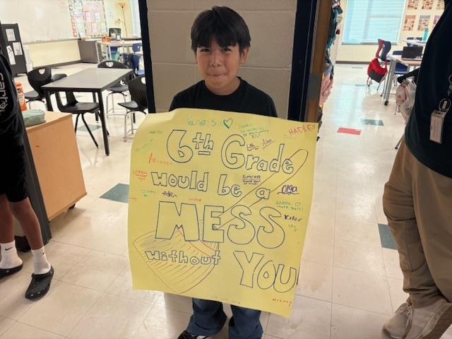A student holding a sign saying, "6th Grade would be a mess without you."