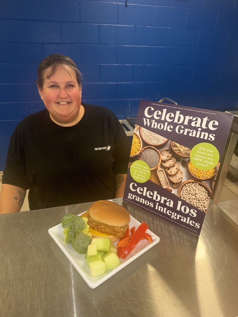 Child Nutrition Staff member standing by a display plate that has a fish sandwich, fruit, and vegetables on it.