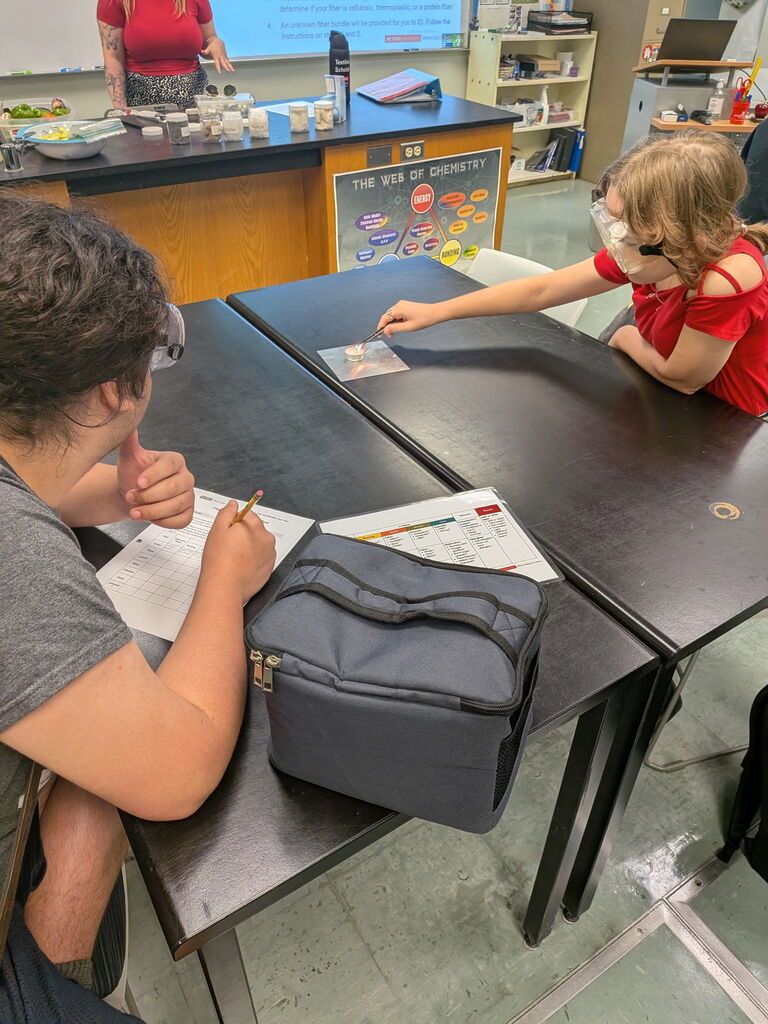 student lights textile sample on fire while their group mate observes and takes notes