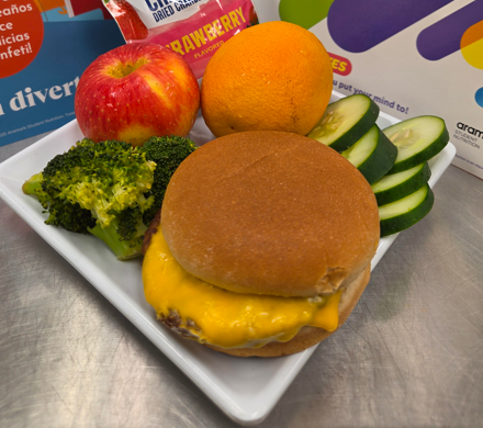 Cheeseburger on plate with broccoli, apple, orange, and cucumbers