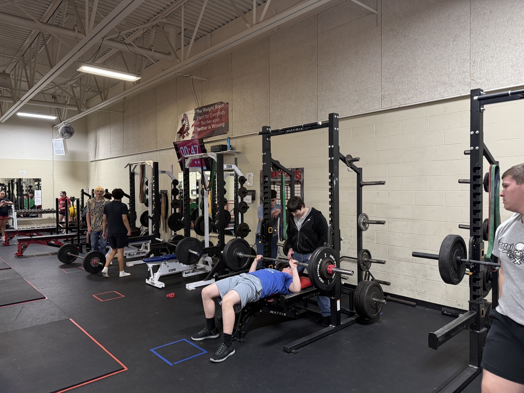 This photo shows a high school–style weight room during what appears to be a strength training class.  In the foreground, a teenage boy is lying on a flat bench performing a barbell bench press inside a black power rack. The barbell is loaded with large black weight plates on both sides. A second student stands closely behind the bench, acting as a spotter and watching the lift carefully. The lifter is wearing a blue T-shirt, gray athletic shorts, black socks, and black training shoes.  To the right edge of the image, another teenage boy stands near a separate rack, facing toward the lifter. He is wearing a light gray T-shirt and appears to be observing or waiting for his turn.  In the background, several other students are scattered around the room. Some are walking between racks, while others stand near benches and weight trees holding stacked plates. There are multiple squat racks and benches lined up in rows, each equipped with barbells and plates. The flooring is black rubber with colored tape squares marking lifting stations.  Mounted high on the back wall is a digital timer displaying “00:47” in bright red numbers, suggesting a timed workout interval. Above it hangs a banner with motivational wording about the weight room. The walls are light-colored cinderblock, and the ceiling is high with exposed white beams and industrial lighting.  Overall, the scene conveys an organized, supervised strength training session with multiple students actively engaged in lifting and moving between stations.