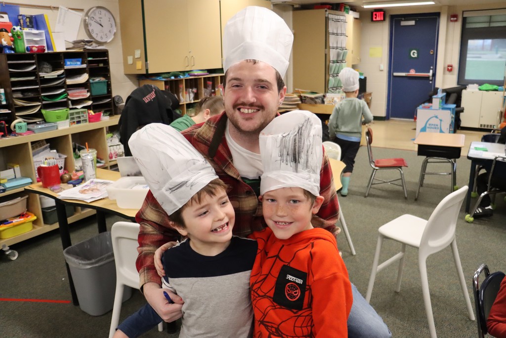 2 Students posing with teacher in classroom, chef hats on their heads