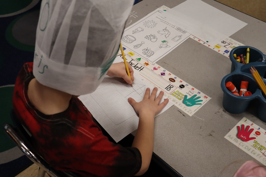 Kid sitting at desk with chefs hat on, drawing a house on paper