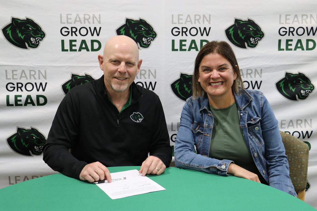 A man and a woman sit at a table in front of a Pennfield Schools backdrop featuring a panther logo and the words “Learn, Grow, Lead.” The man signs a document while smiling at the camera, and the woman sits beside him, also smiling.