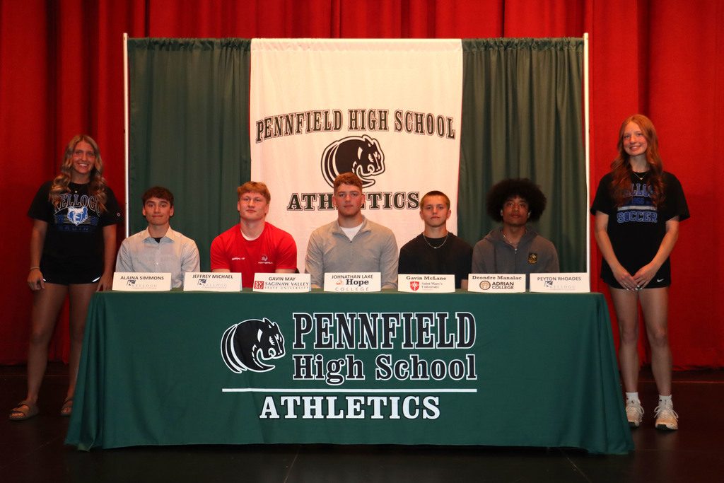 Seven Pennfield High School student-athletes pose at a signing day table in front of a “Pennfield High School Athletics” backdrop. Six students are seated behind a table with name signs showing their names and colleges, while one student stands to the right. A female student also stands on the far left. The athletes are dressed in a mix of school apparel and casual clothing and are being recognized for committing to play college sports.
