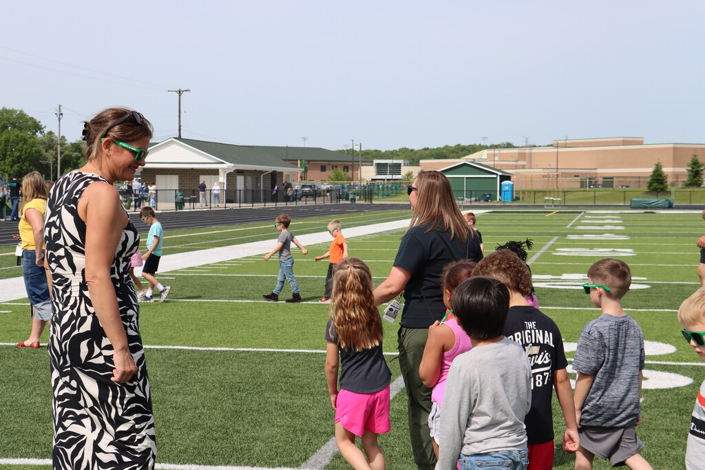 Students and staff wearing green sunglasses walk together on the football field during a Pennfield end-of-year celebration. Children are gathered in small groups on the turf while staff members look on. School buildings and athletic facilities can be seen in the background on a sunny day, marking the start of summer break.