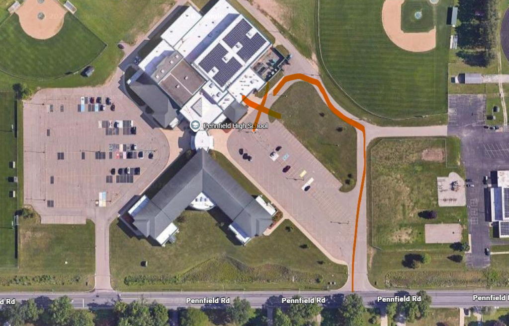 Aerial map of the Pennfield High School campus showing the designated traffic route for summer meal pickup. An orange line begins at the entrance from Pennfield Road on the east side of the property, continues north through the main parking lot, loops around the front of the high school entrance, and exits near the school entrance area. The route is highlighted to direct vehicles through the pickup location. School buildings, parking lots, athletic fields, and Pennfield Road are visible in the satellite image.