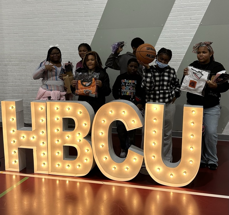 7 students showcasing basketballs, clothing items, drinking cups and more that they won during the fair standing behind the illuminated HBCU letters   
