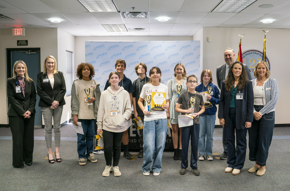 Group photo of students and staff standing in a district office space, holding certificates and trophies during an academic recognition event, with a Peoria Unified School District backdrop and flags visible behind them.