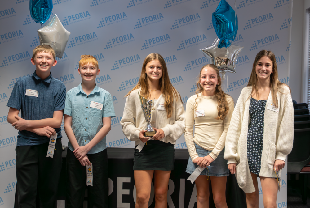 Five students stand in a row holding awards and ribbons in front of a Peoria Unified School District backdrop. Blue and silver star-shaped balloons are displayed behind them, and a table with the Peoria logo is visible in front.