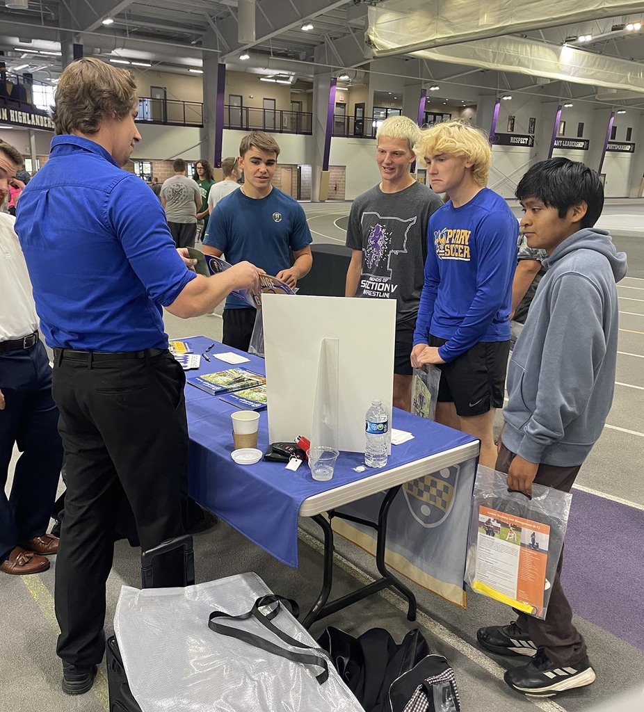 Several high school students gathered around a table while an adult male is speaking.