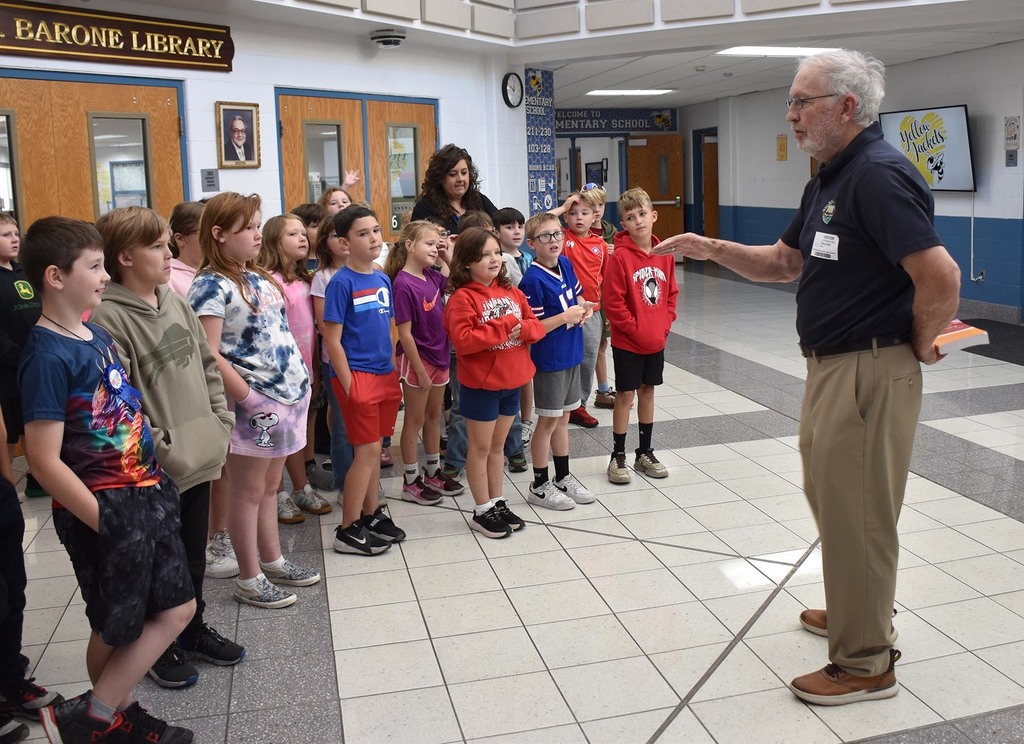 An adult male talking to a group of 3rd graders in the main hallway of a school. 