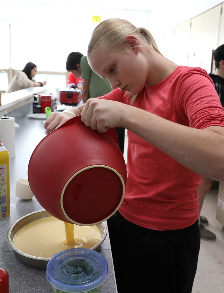 A jr. high girl pouring a mix into a pan. 