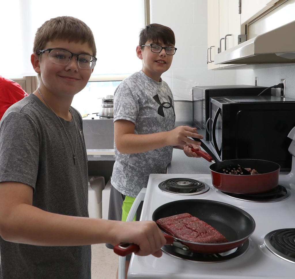 Two jr. high males cooking meat on a stove. 
