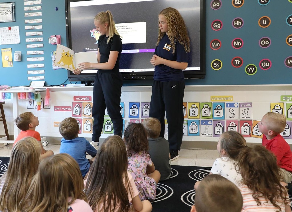 Two 8th grade girls reading a book to kindergartners. 