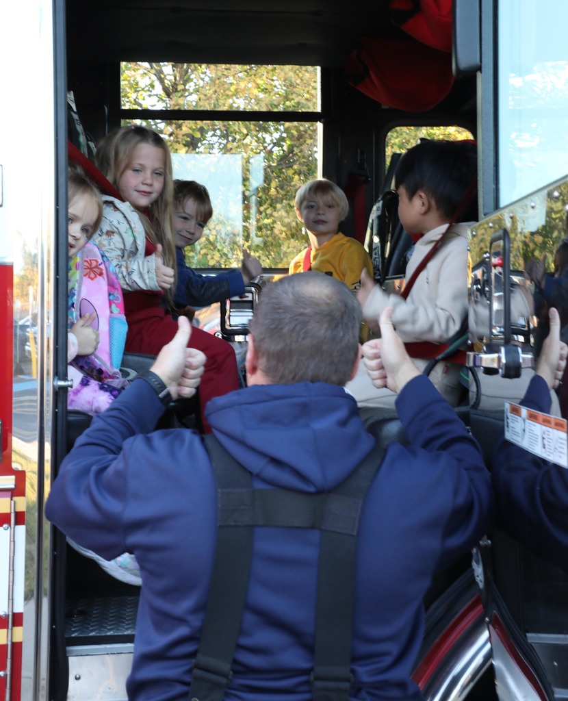 Kids sitting in a fire truck giving thumbs up to a fireman. 