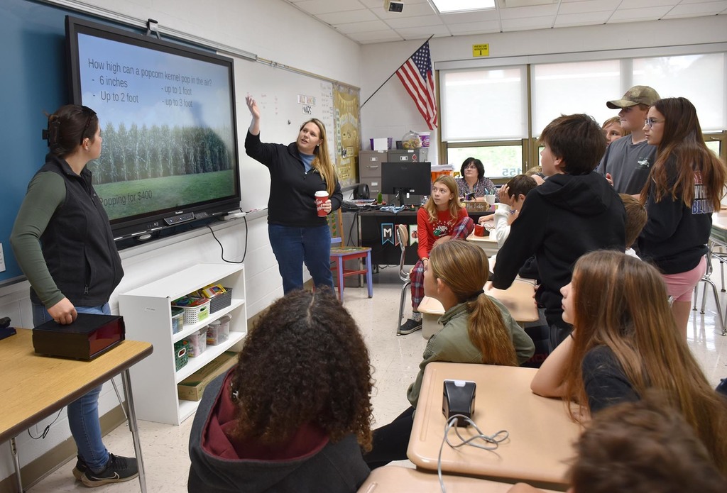 Two teachers standing at a smart board as a group of students looks on. 