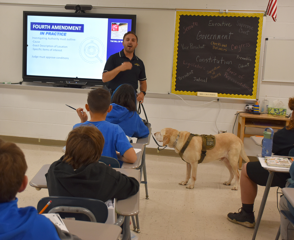 An adult male, holding onto a dog on a leash, stands in front of students in a classroom. 
