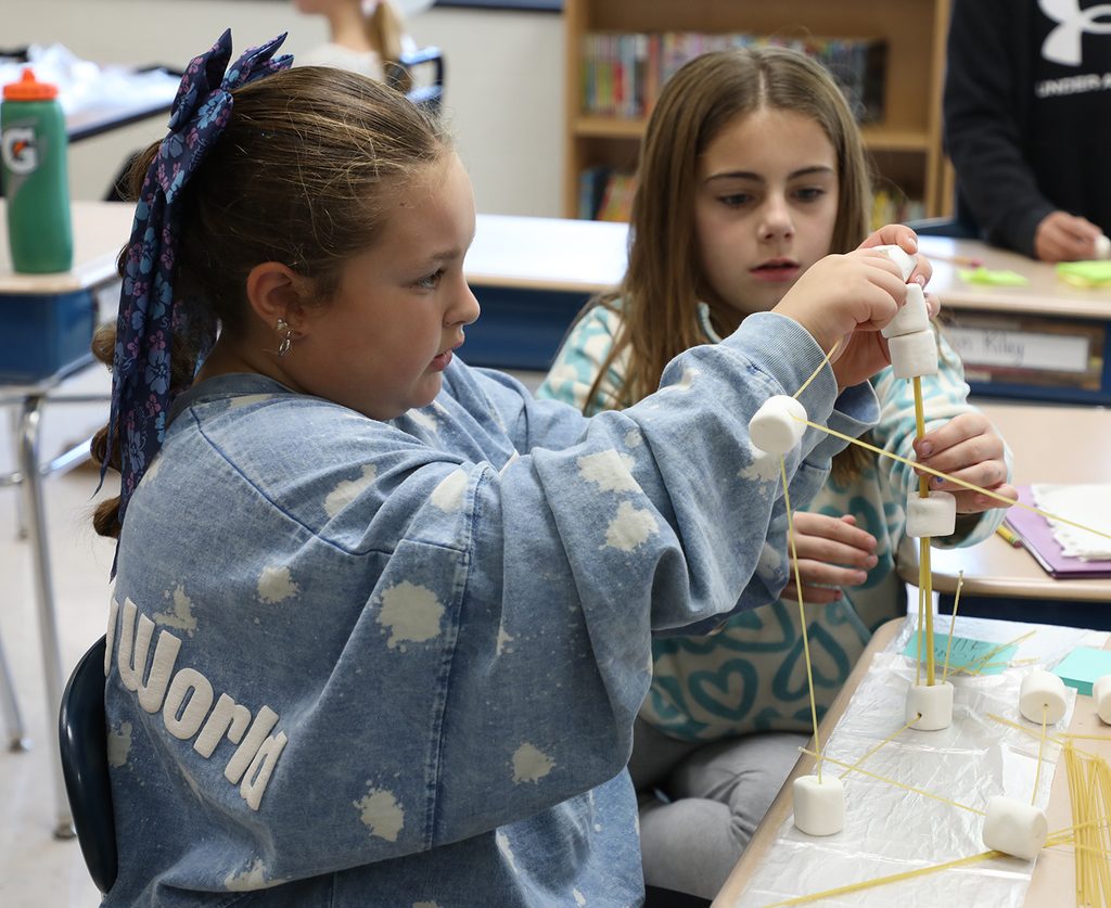 Two elementary girls trying to build a tower with marshmallows and uncooked spaghetti. 