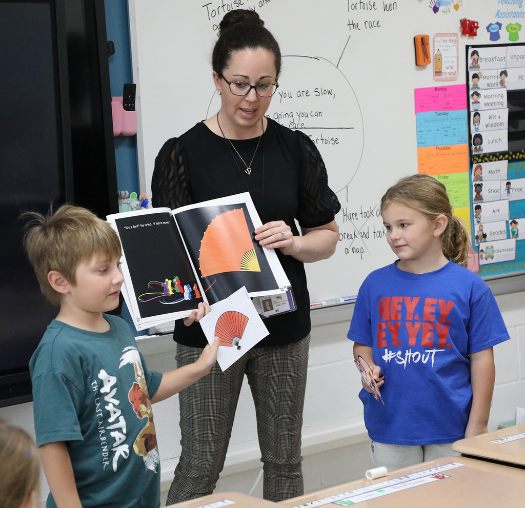 A female teacher holding a book while two elementary students stand next to her. 