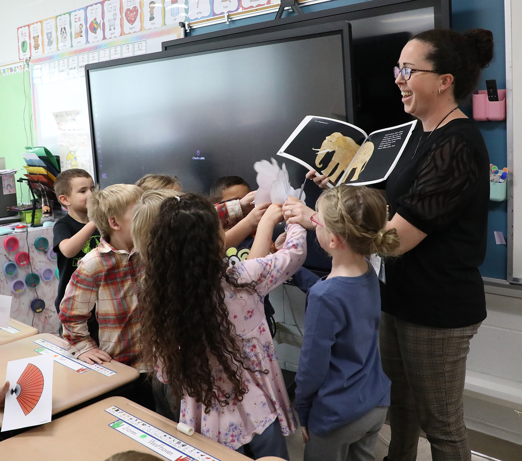 A teacher, holding a book, surrounded by elementary students. 