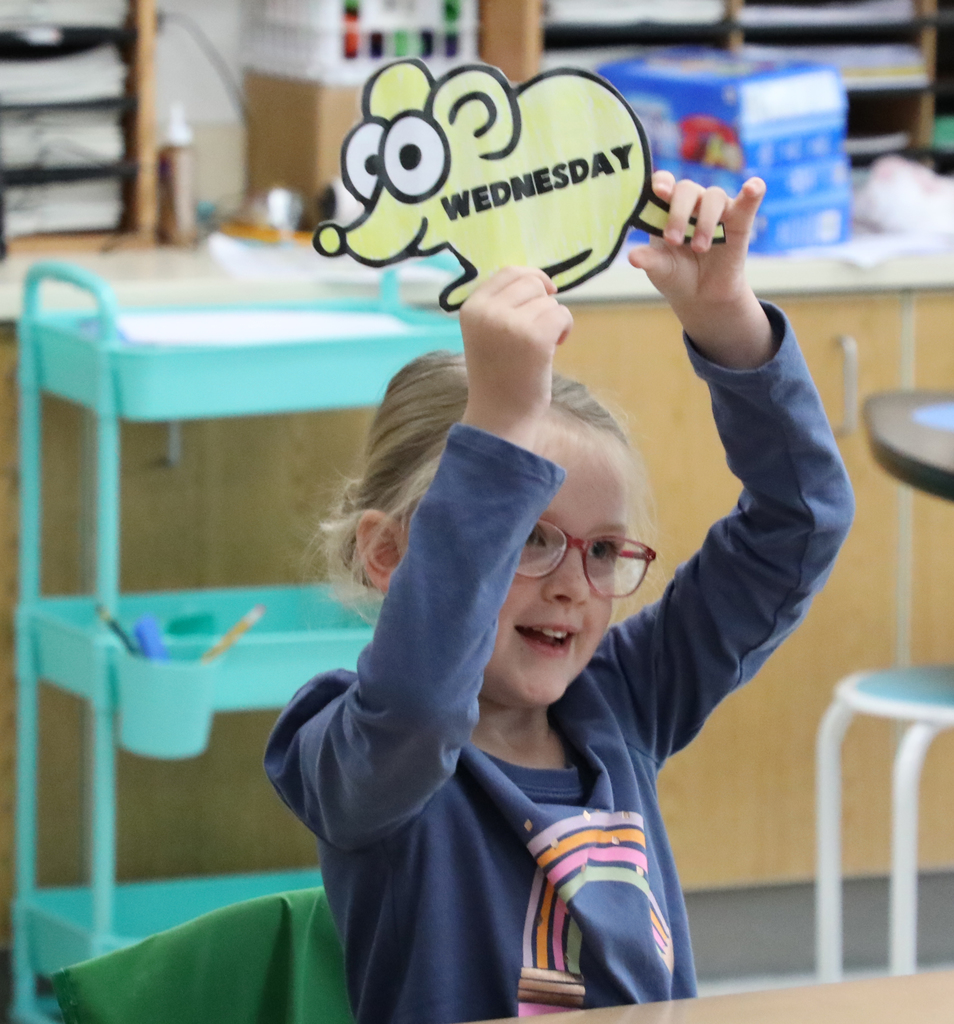 An elementary girl holding up a yellow paper mouse with the word "Wednesday" on it. 