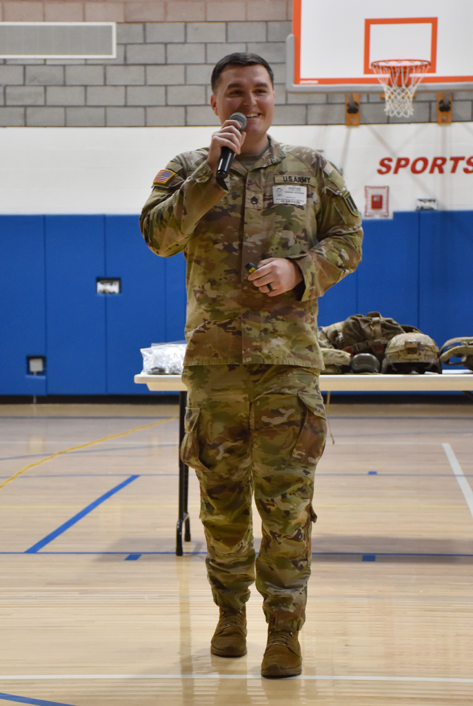 A male, holding a microphone, standing in a school gym. He is wearing military attire. 
